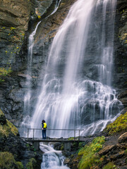 Espectacular cascada en un río de alta montaña con un puente que atraviesa el cauce y un montañero con mochila amarilla en primer plano
