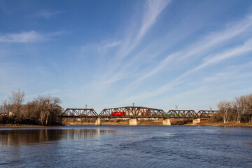 Fototapeta premium Train crossing a bridge over a body of water with blue skies and soft clouds in the background, Winnipeg, Manitoba