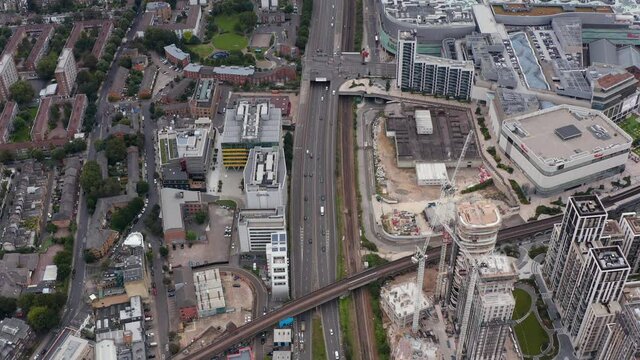 Aerial view of busy multilane road leading along construction site of development project. Tilt down footage to railway bridge. London, UK