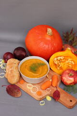 Still life photo with squash, red apples, autumn leaves, onion, carrot, fresh bread, bowl of pumpkin soup on a table. Gray background with copy space. Comfort food concept. Thanksgiving menu ideas. 