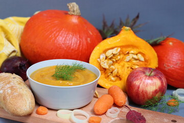 Still life photo with squash, red apples, autumn leaves, onion, carrot, fresh bread, bowl of pumpkin soup on a table. Gray background with copy space. Comfort food concept. Thanksgiving menu ideas. 