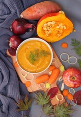 Seasonal vegetables, creamy pumpkin soup and autumn leaves on grey background. Colorful picture of pumpkin soup ingredients on a table. Comfort food concept. 