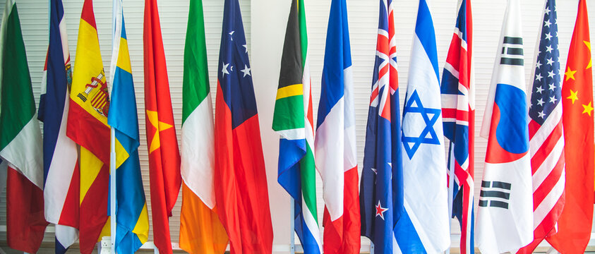 Closeup of a collection of various flags of different countries standing tall together in a row on a stand against white background