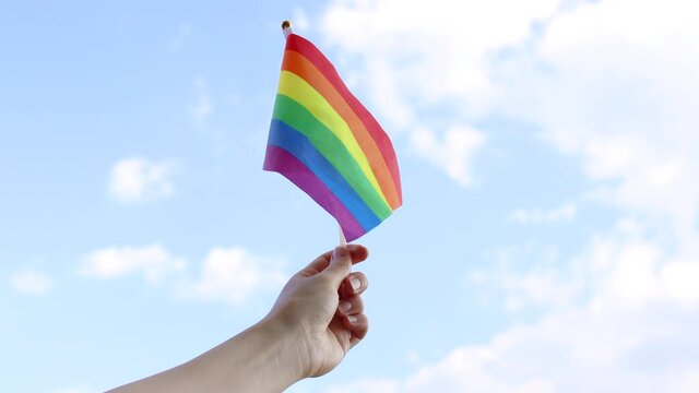 Girl hand waving a small colorful rainbow gay pride flag against a light blue cloudy background