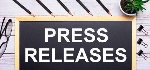 On a light wooden table - a board with the text PRESS RELEASES pencils, plants, glasses and paper clips. Business concept