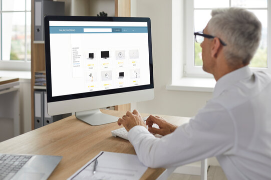 Man shopping online. Senior businessman sitting at work desk, looking at computer display, browsing Internet gadget outlet, buying new office equipment, adding items to cart, paying with credit card