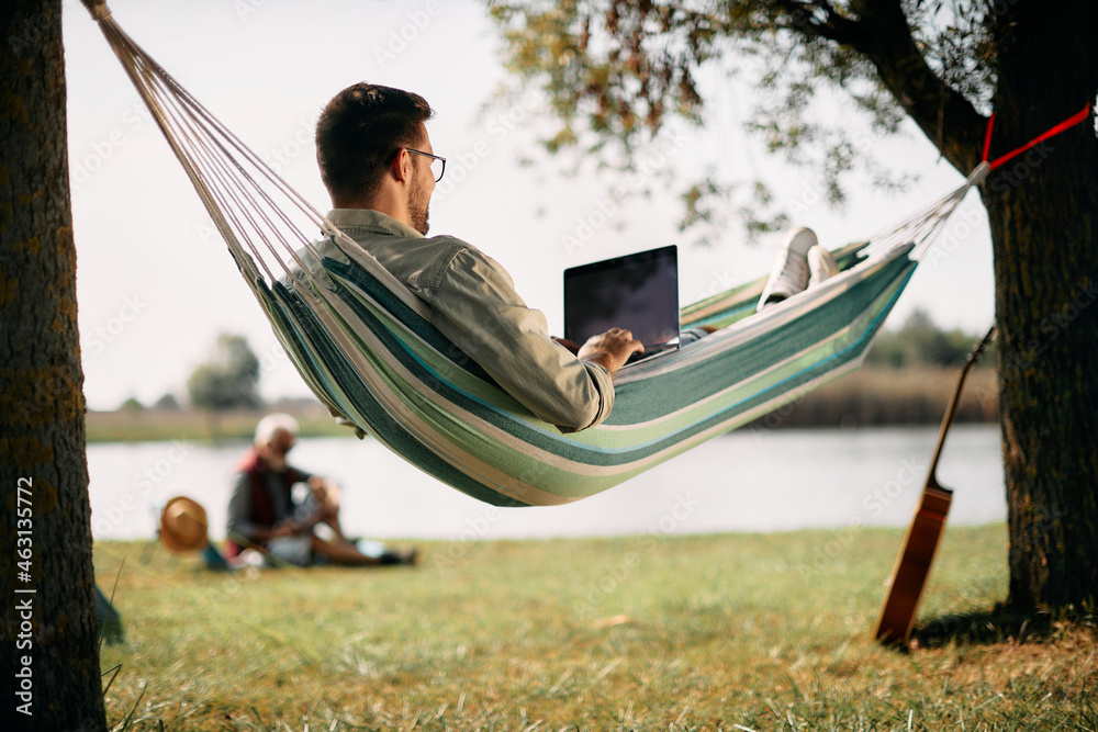 Wall mural below view of man works on laptop while relaxing in hammock by lake.