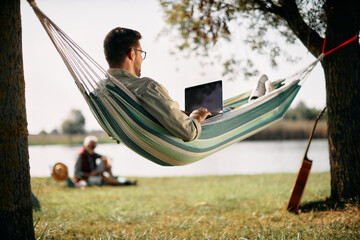 Below view of man works on laptop while relaxing in hammock by lake.