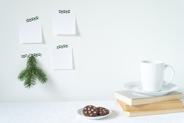 Festive breakfast still life.Cup of coffee with cookies, books on desk.Fir branch, empty notepads mockup hanging on the wall.Christmas decor.Working space, home office.Happy new year resolutions, list