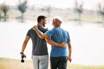 Rear view of athletic father and son talk while walking embraced by the lake.
