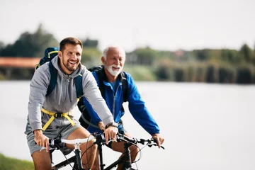 Poster Im Rahmen Happy athletic man enjoys in cycling with his senior father by the river. Copy space. © Drazen