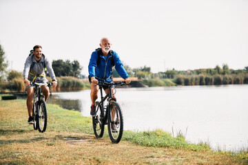 Happy senior athlete and his son ride bicycles by the lake in nature. Copy space. - Powered by Adobe