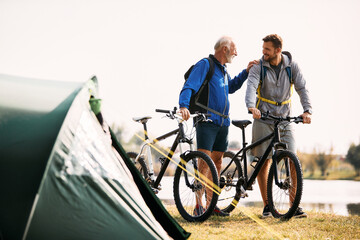 Happy mature father and son talk while getting ready for cycling during their camping day.