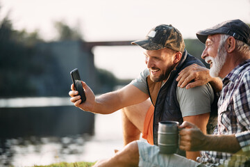 Happy man and his senior father take selfie with smartphone while camping by lake.
