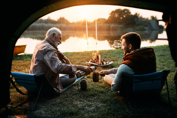 Senior fisherman and his son enjoy in hot drink while camping by water at sunset.