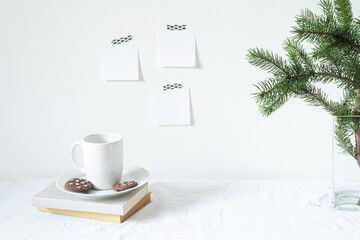 Obraz na płótnie Canvas Festive breakfast still life.Cup of coffee with cookies, books and vase with fir branch.Empty notepads mockup hanging on the wall.Christmas decor.Working space, home office.Happy new year resolutions
