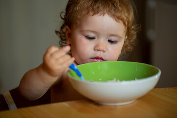 Portrait of funny little baby boy eating from plate holding spoon closeup.