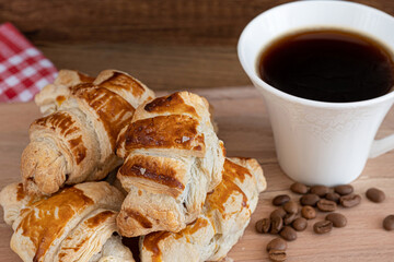 Crispy croissants presentation with cup of hot filter coffee and beans on wooden plate. Fresh brown tasty croissants on table, popular breakfast or delicious lunch ingredients in French culture. 