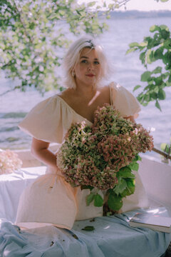 A blonde girl of European appearance is smiling, sitting in a wooden boat filled with flowers in a light dress, holding a bouquet of hydrangeas in her hands