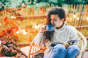 Young woman leafs through book and takes warm beverage cup sitting in armchair on balcony decorated with yellow lights and autumn attributes in evening