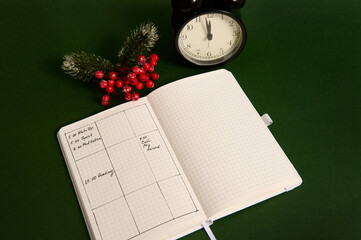 Fototapeta na wymiar Top view of an opened page of notepad with plans for day, alarm clock and snowy branch of holly Christmas plant isolated on green background