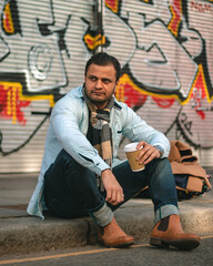Muscular man sitting on the sidewalk having coffee against graffiti background