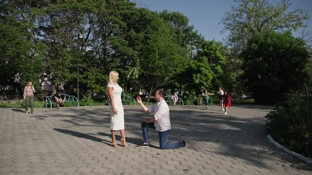 young man in love is on his knees and makes an offer with help of ring to his beloved woman, crowd of people runs to couple, shouting no in park background of trees