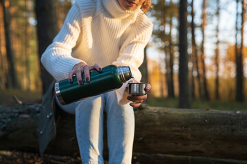 Young woman in cozy sweater pour hot tea from thermos in cup walking in autumn forest. Female enjoy fall nature warming up with steaming beverage. Girl taking break from hike wondering in fall woods
