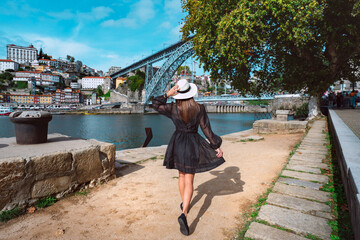 Young woman tourist in black dress enjoying beautiful view of Porto city and famous famous Dom Luis I Bridge. Summer holiday vacation in North Portugal. - obrazy, fototapety, plakaty
