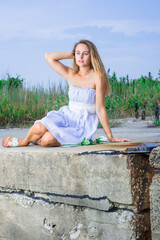 A pretty blonde girl, dressing in a white blue dress, white waistband and sandals,  is sitting on a high land, thoughtfully looking forward. A white rose is put on the ground..