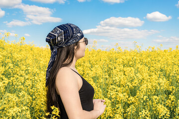 A girl in sunglasses and a bandana looks at a blooming rapeseed field
