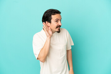 Young caucasian man isolated on blue background listening to something by putting hand on the ear