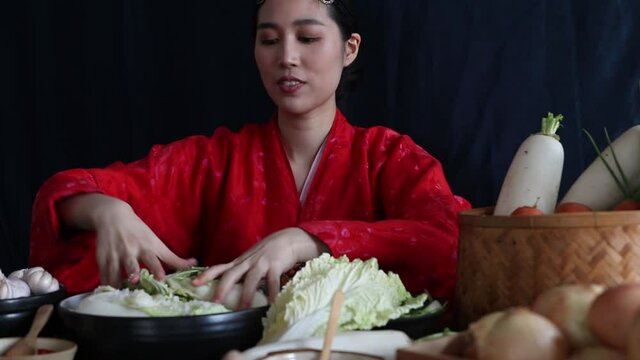 Beautiful asian woman dressed in red hanbok makes kimchi, a traditional Korean dish, sprinkles salt in a bowl of white cabbage, and she meticulously kneads it with clean, fresh vegetables.