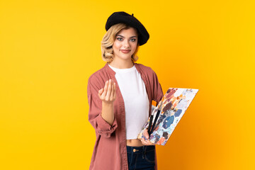 Young artist girl holding a palette isolated on yellow background inviting to come
