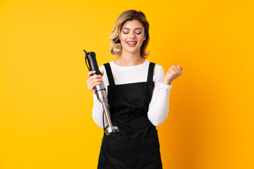Girl using hand blender isolated on yellow background celebrating a victory in winner position