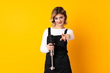 Girl using hand blender isolated on yellow background pointing front with happy expression