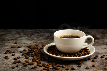 Close-up of hot coffee in a white cup is placed on cement floor table, lots of roasted coffee beans are in coffee cup saucer, and around, smoke and aroma waft from the cup. Black, Blurred background