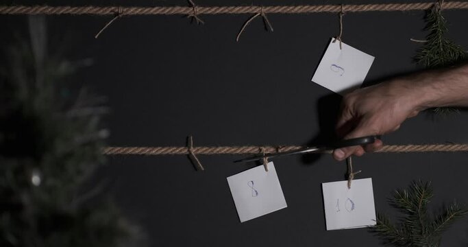 homemade DIY advent calendar, created on a black background and tied with a craft string, in the foreground is a Christmas tree with a garland. scissors extend to cut one of the tags