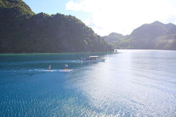 Fototapeta na wymiar aerial view of people paddling sup standup paddle boards in calm waters with a Bangka boat and beaches and mountains in the background. Philippines