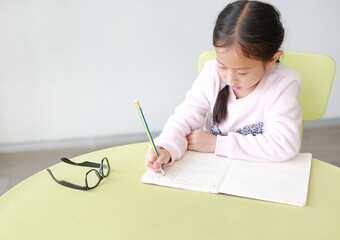 Portraits of little Asian girl writes in a book or notebook with pencil sitting on kid chair and table in classroom.