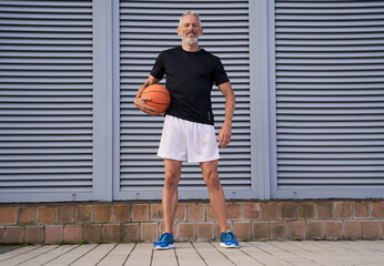 Active middle aged man in sportswear holding basketball ball and smiling at camera while standing outdoors over gray background