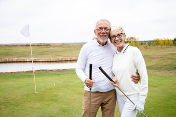 Portrait of smiling caucasian senior couple holding golf clubs and standing on the course.