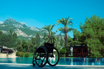 an empty black wheelchair stands by the pool against the backdrop of palm trees and blue sky,...