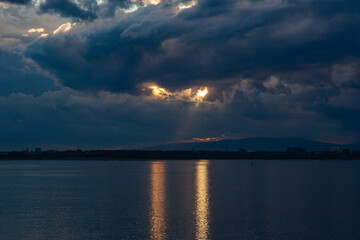 The sky is covered with clouds at sunset in the sea bay, two beams of light make their way through the clouds and beat off the reflection in the water.
