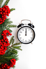 New Year and Christmas card. Black vintage alarm clock on a white background with sequins. Green Christmas tree and pine with red rowan berries. Holiday wallpaper.