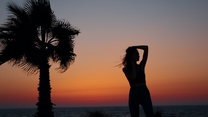 Sporty slim woman in silhouette standing on ocean beach after workout and enjoying beautiful colorful sunset. Evening training on fresh air. Active hobby concept.