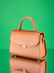Fashionable handbag made of reddish-beige leather, standing on a mirror surface, on a rich green background with a reflection. Copy space.
