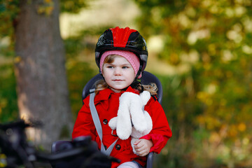 Little toddler girl with security helmet on head sitting in bike seat of her mother or father bicycle. Safe and child protection concept. Family ctivity trip on autumn day. Kid holding push soft toy