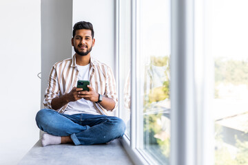 Handsome indian young man sits home on windowsill, reads text messages, sends email, uses his phone and fast internet connection.