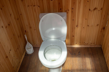 white toilet with plastic lid in wooden outdoor toilet. Plastic brush in background